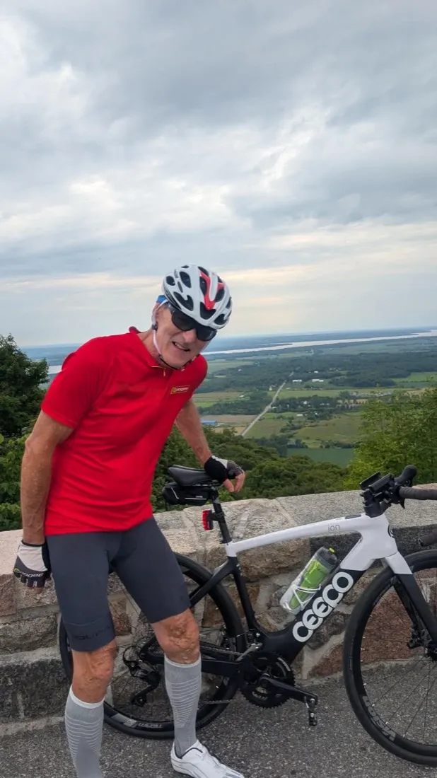Kirk Douglas in a red jersey and white helmet standing with a CEEPO e-bike at a stone lookout wall in Gatineau Park, farmland and the Ottawa River far below behind him.